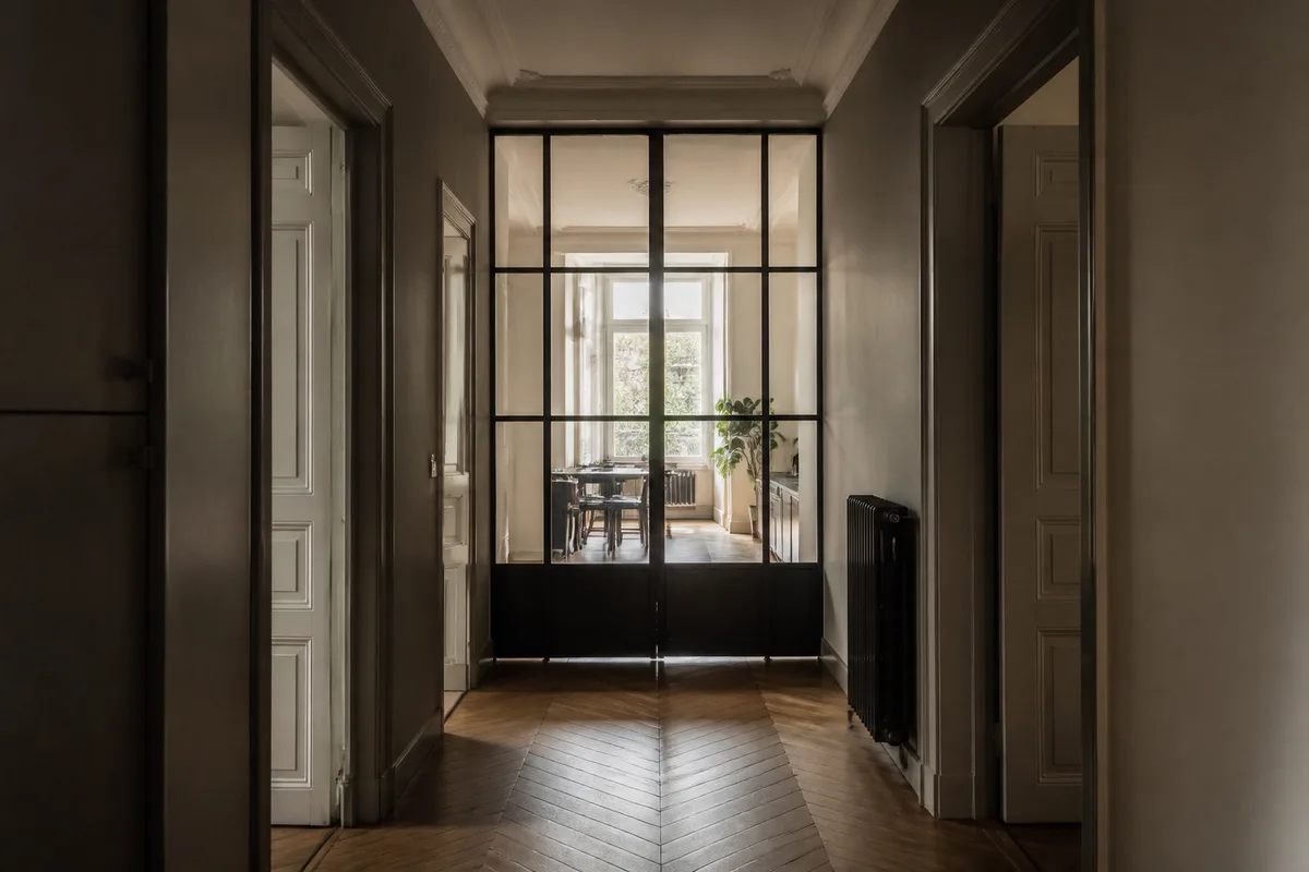 Cloison vitrée en acier noir séparant une cuisine d’un séjour dans un logement ancien, lumière traversante de fin de matinée