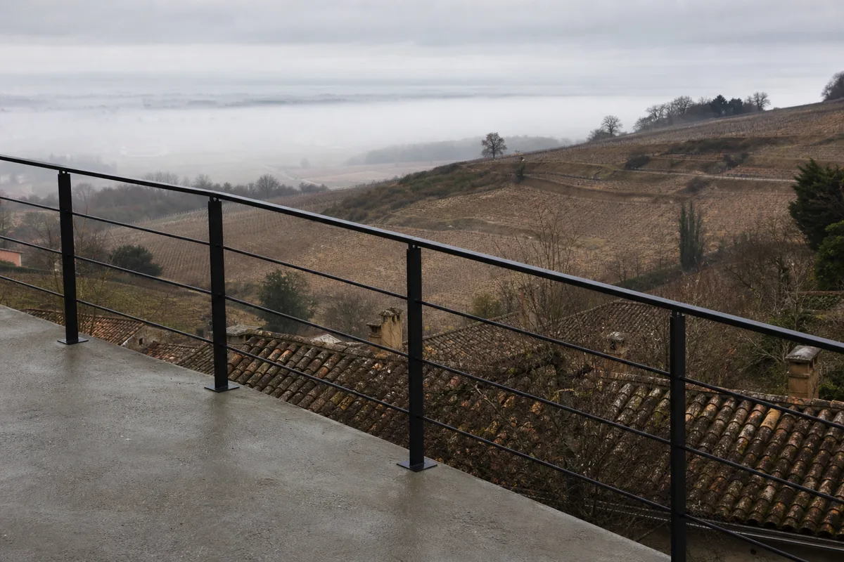 Garde-corps acier noir mat suivant la pente d’une terrasse dominant les coteaux, lumière grise de fin d’hiver